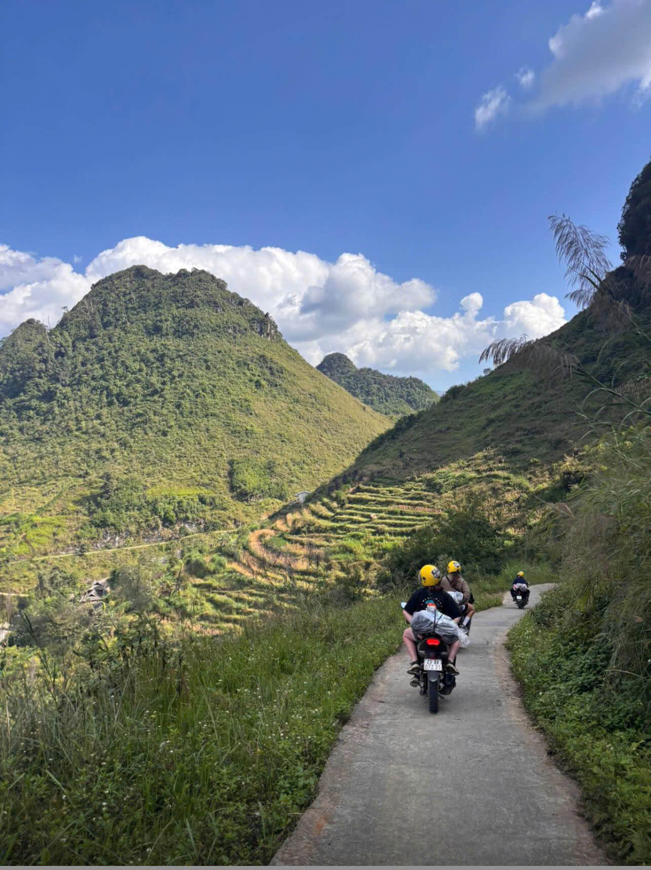 Motorbike riders with yellow helmets navigating a narrow concrete path through lush green terrace mountains, showing the authentic rural routes of a Ha Giang pickup service.